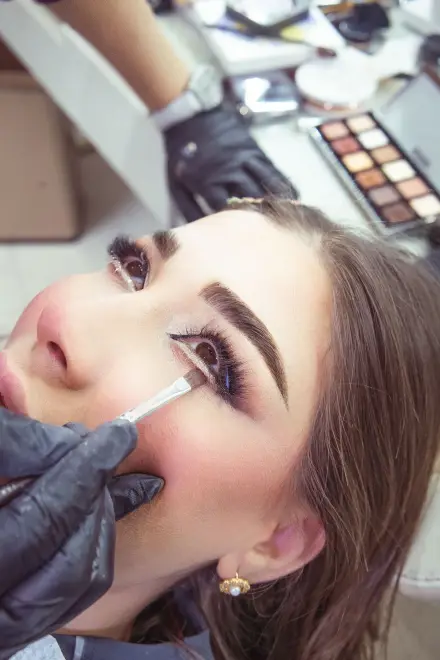 Maquilladora delineando los ojos de una clienta, con la paleta de sombras detrás
